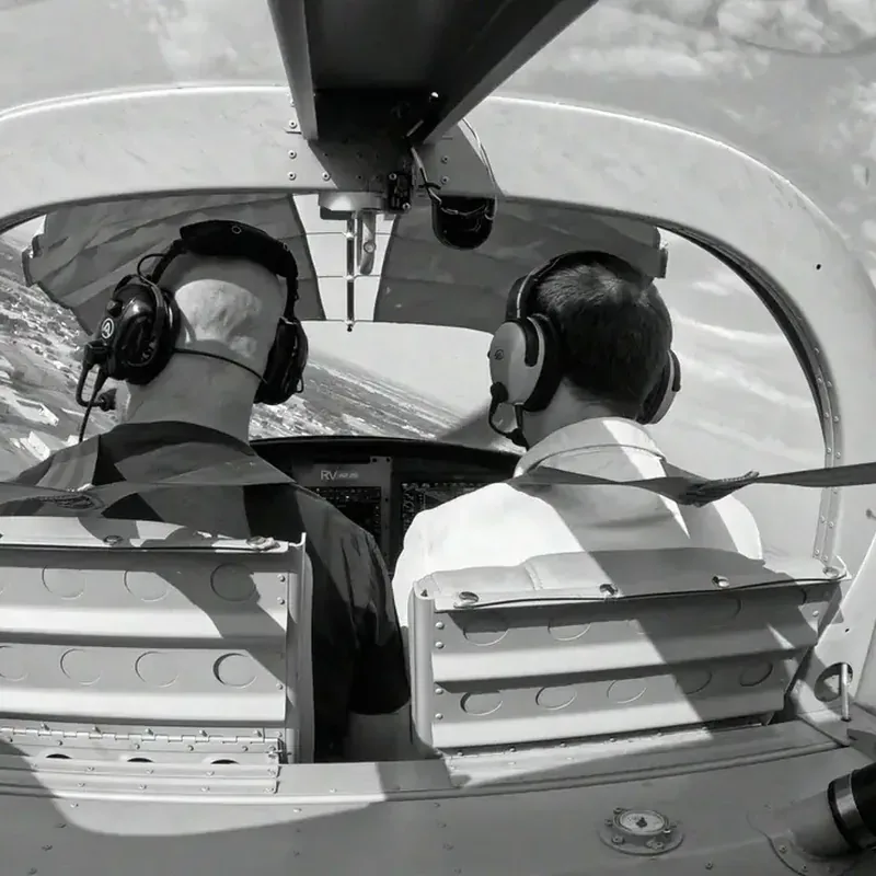 Black and white photo of two pilots in headsets seen from the back seat, flying over open country