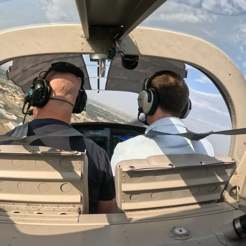 Two pilots in headsets seen from the back seat, flying over open country